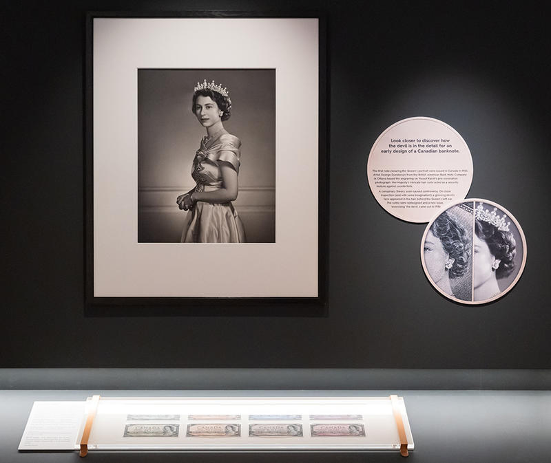 Money Talks exhibition gallery display of Queen Elizabeth II's portrait next to the a photo showing the devil in the hair Money Talks exhibition gallery display of Queen Elizabeth II's portrait next to the a photo showing the devil in the hair pointed out and a display of the Canadian banknotes below